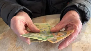 Două mâini cu pielea ridată țin mai multe bancnote de culoare verde și galbenă deasupra unei suprafețe plane și deschise la culoare.