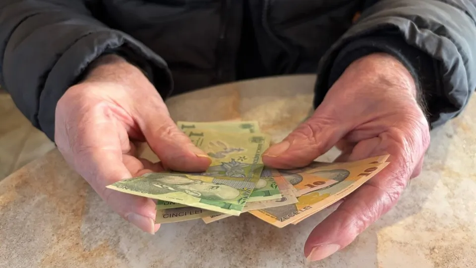 Două mâini cu pielea ridată țin mai multe bancnote de culoare verde și galbenă deasupra unei suprafețe plane și deschise la culoare.
