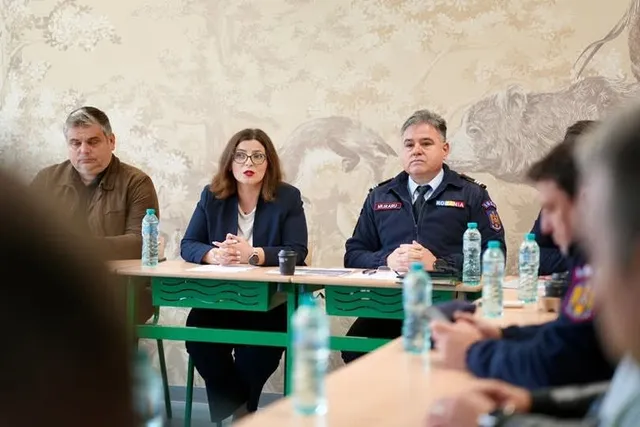 O femeie cu ochelari și un bărbat în uniformă stau la o masă cu sticle de apă în fața unui perete tapetat.