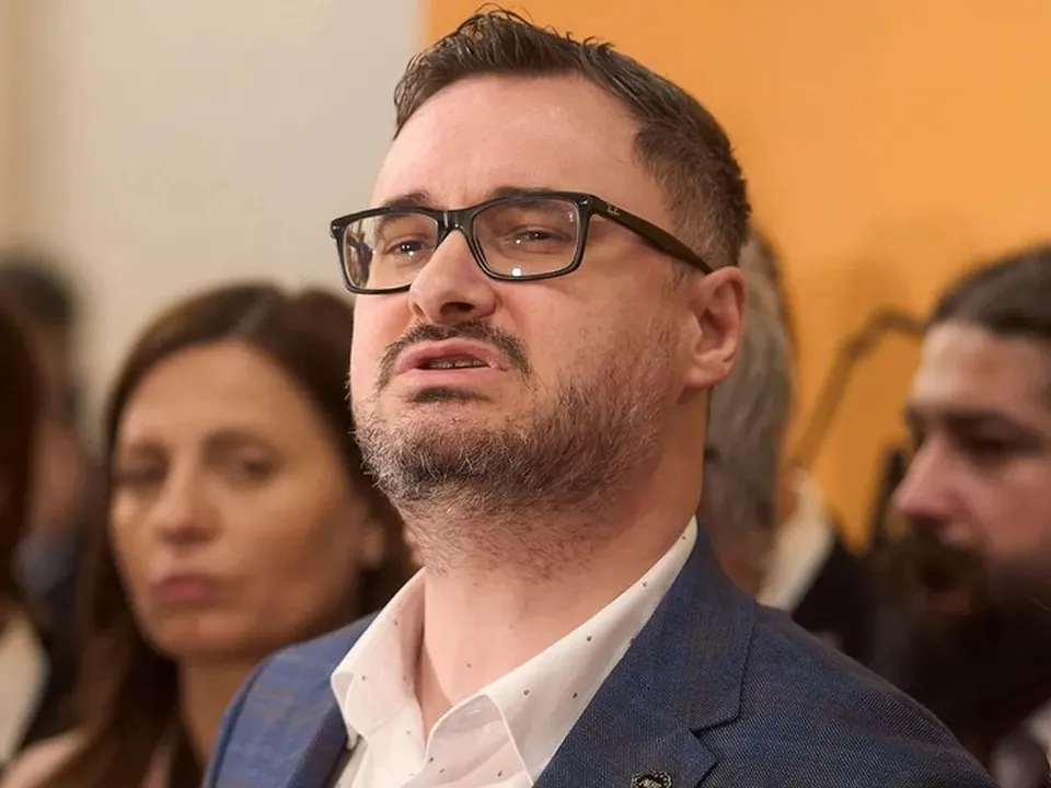 Un bărbat cu ochelari și barbă scurtă poartă o cămașă albă cu puncte și un sacou albastru în fața unor persoane din fundal.