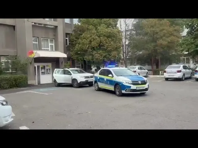 O mașină de poliție albastră și albă este parcată pe un teren asfaltat lângă alte vehicule albe și un bloc de apartamente.