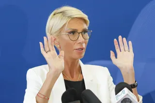 O femeie cu părul blond, purtând ochelari și o jachetă albă, ridică ambele mâini în poziție laterală în fața unor microfoane.