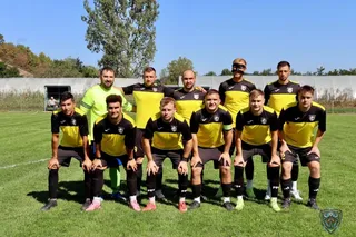 O echipă de fotbaliști în echipamente galbene și negre pozează pe un teren de fotbal sub un cer senin.