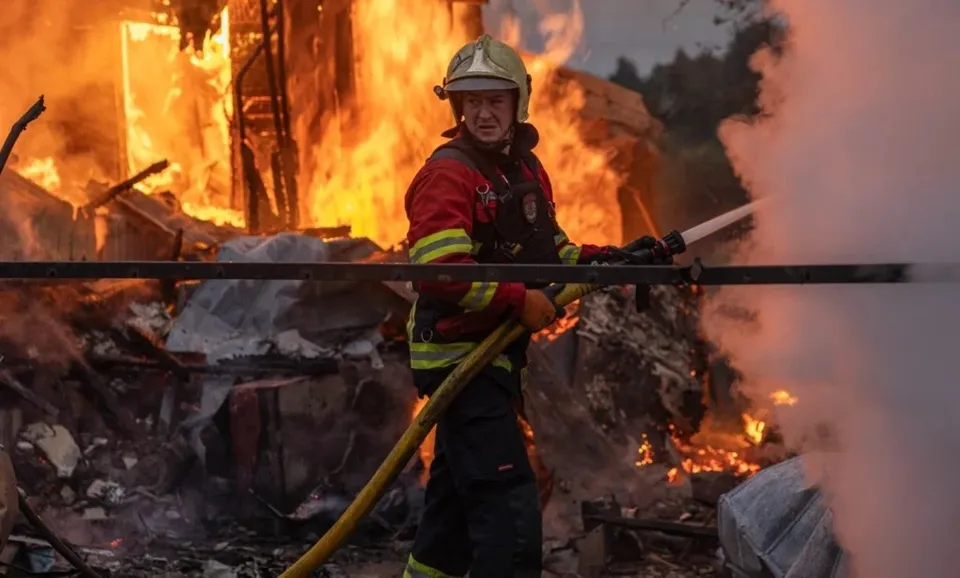 Un pompier cu echipament de protecție roșu și negru stă lângă un incendiu de structură și ține un furtun de apă.