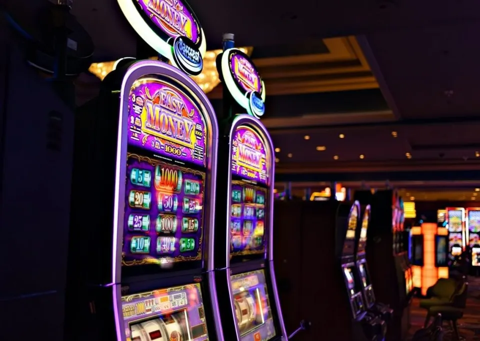 Mai multe aparate de joc de tip slot machine sunt aliniate într-o încăpere întunecată cu lumini colorate.