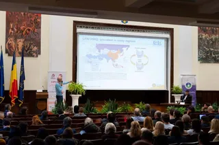 Un bărbat stă în picioare lângă un ecran de proiecție mare și prezintă o hartă unei mulțimi de oameni așezate în auditorium.
