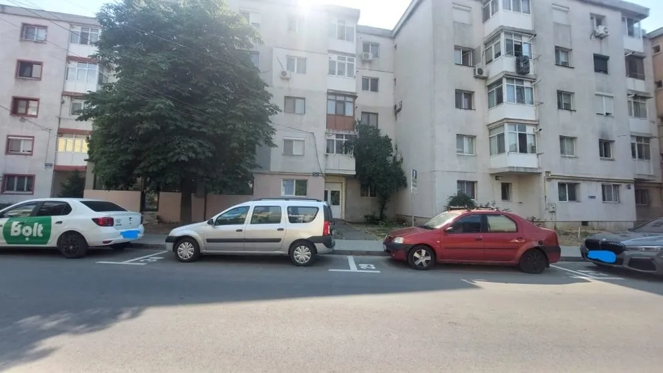 Mai multe mașini sunt parcate pe un loc de parcare marcat cu linii albe în fața unor blocuri de apartamente.