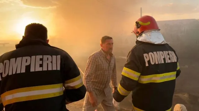Doi pompieri cu spatele, în echipament, și un bărbat în cămașă în carouri stau în fața fumului dens și a unei lumini portocalii.