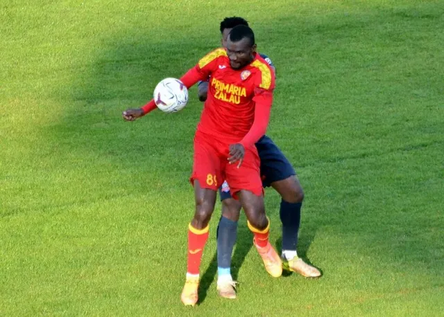 Un fotbalist în echipament roșu controlează mingea pe teren, în timp ce un adversar în albastru îl urmărește.