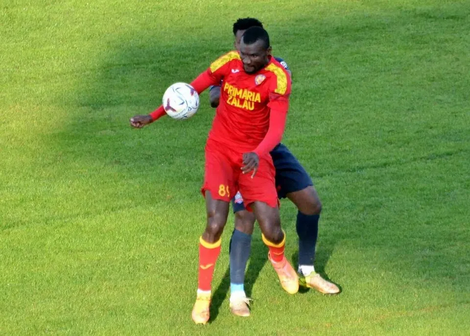 Un fotbalist în echipament roșu controlează mingea pe teren, în timp ce un adversar în albastru îl urmărește.