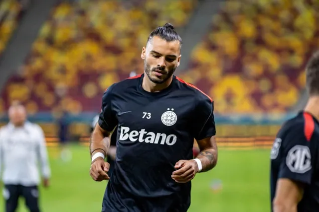 Un bărbat cu părul strâns într-un coc și barbă poartă un tricou negru cu inscripția Betano și numărul 73 pe un teren de fotbal.
