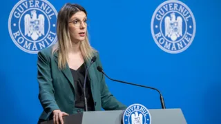 O femeie cu ochelari și sacou verde stă în spatele unui podium gri în fața unui fundal albastru cu sigilii circulare.