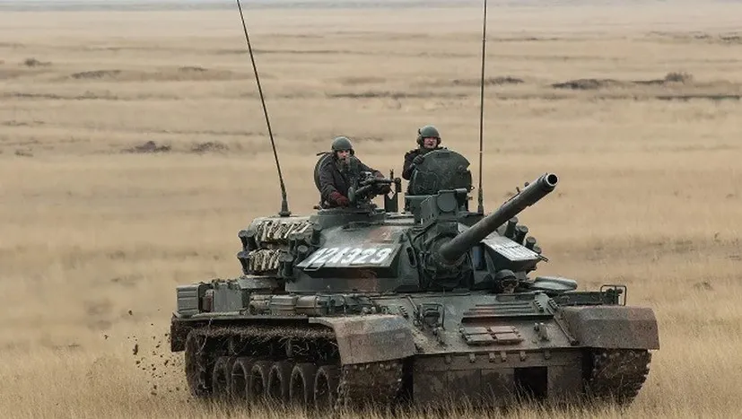 Un blindat cu un tun și doi militari în partea superioară se deplasează pe un teren deschis cu iarbă uscată.