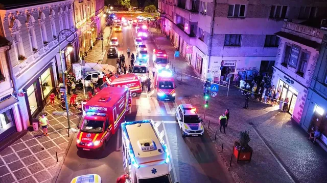 Mai multe ambulanțe și mașini de poliție cu luminile aprinse sunt parcate pe un drum urban între clădiri în timpul nopții.