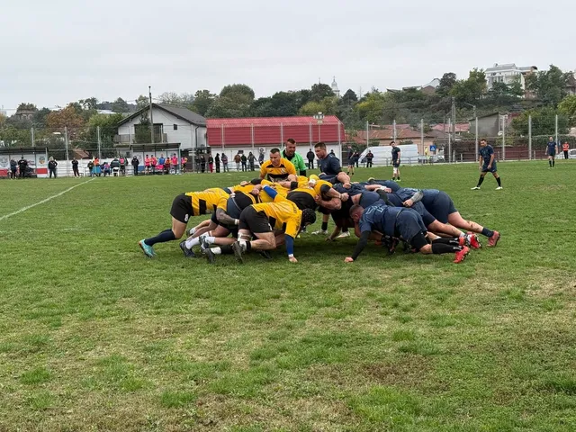 O grupă de jucători de rugby îmbrăcați în galben și albastru formează un scrum pe un teren de iarbă verde.