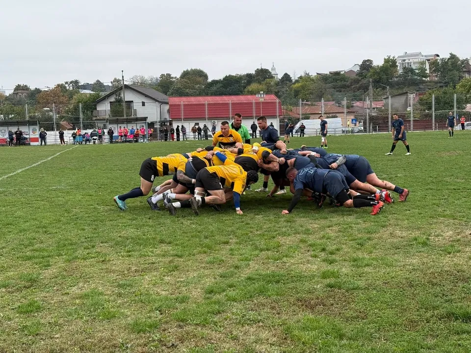 O grupă de jucători de rugby îmbrăcați în galben și albastru formează un scrum pe un teren de iarbă verde.