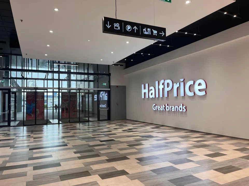Un semn suspendat cu pictograme negre atârnă deasupra unui hol cu podea modelată, în fața unui perete alb cu inscripția HalfPrice.