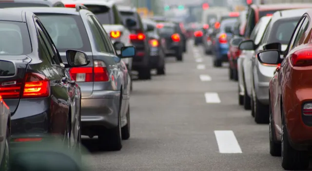 Mai multe mașini sunt aliniate în două rânduri pe o bandă de rulare, formând un trafic dens pe un drum asfaltat.