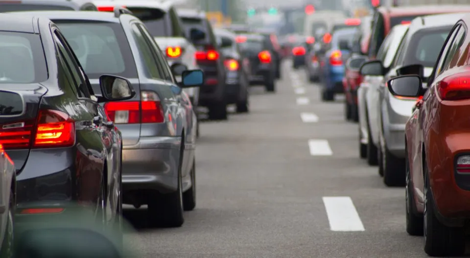 Mai multe mașini sunt aliniate în două rânduri pe o bandă de rulare, formând un trafic dens pe un drum asfaltat.