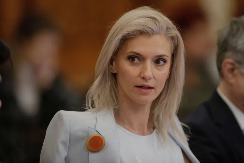 O femeie cu părul blond deschis poartă o vestă albă cu un accesoriu circular portocaliu pe lapel.