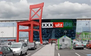 O sculptură gigantă în formă de scaun roșu stă în fața unui magazin de mobilă cu parcarea plină de mașini în prim-plan.