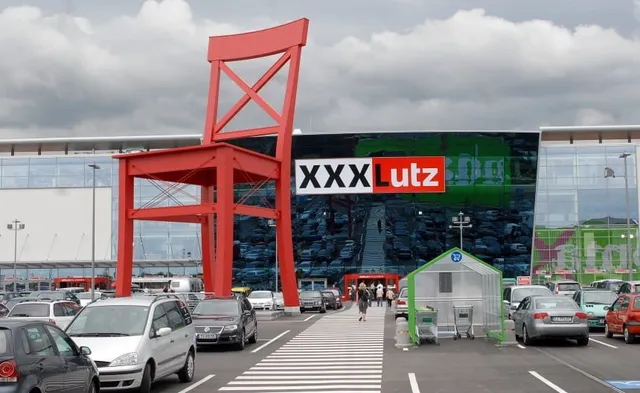 O sculptură gigantă în formă de scaun roșu stă în fața unui magazin de mobilă cu parcarea plină de mașini în prim-plan.