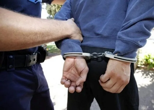 Un agent în uniformă albastră ține de brațul unui bărbat care poartă un hanorac albastru și este înlănțuit cu cătușe la spate.