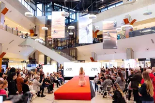 O persoană merge pe o rampă roșie în fața unui public așezat în interiorul unui centru comercial iluminat.