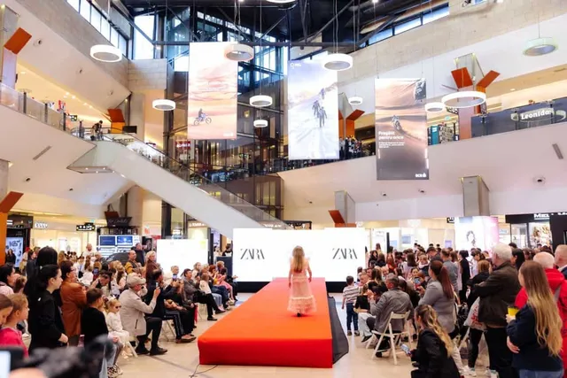 O persoană merge pe o rampă roșie în fața unui public așezat în interiorul unui centru comercial iluminat.