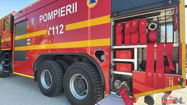 Un camion de pompieri roșu cu inscripții albe este parcat pe un drum, afișând echipamente de intervenție în compartimentul lateral deschis.