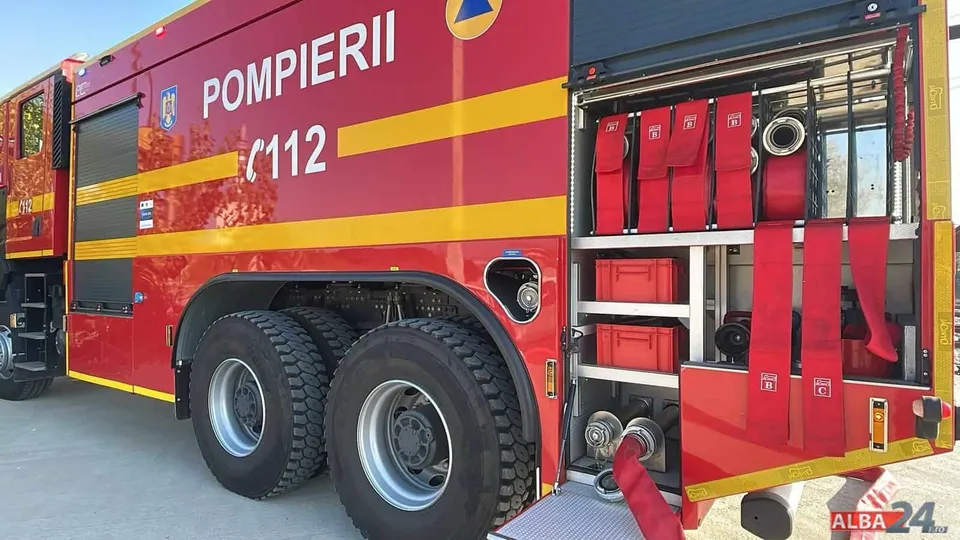 Un camion de pompieri roșu cu inscripții albe este parcat pe un drum, afișând echipamente de intervenție în compartimentul lateral deschis.