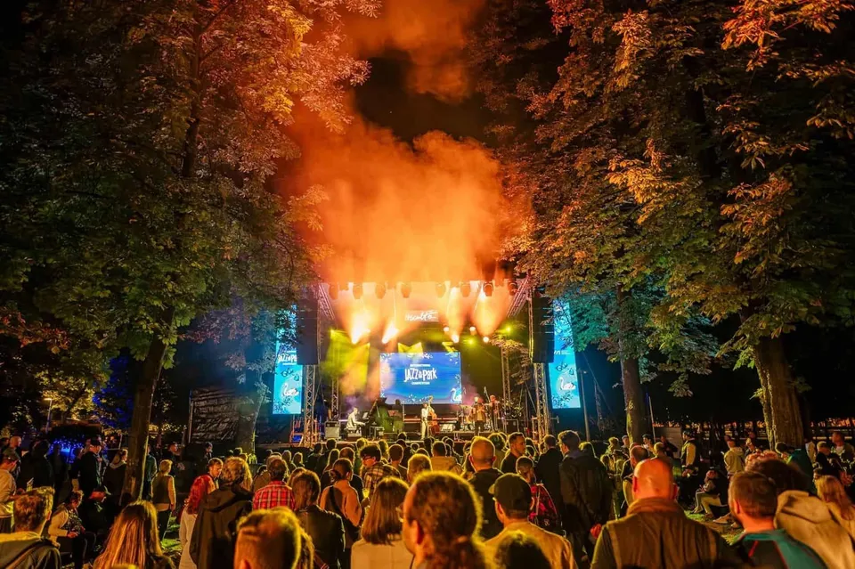 O mulțime de oameni stă în fața unei scene luminate, înconjurată de copaci, la un concert în aer liber.