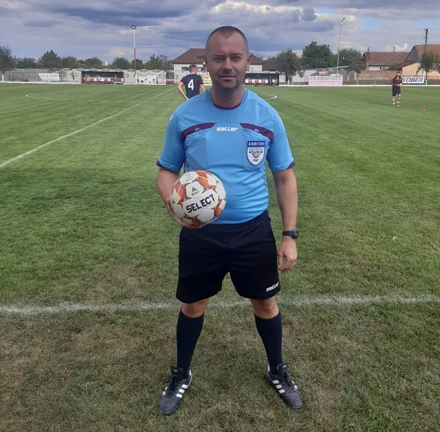 Un bărbat în echipament sportiv albastru și negru stă pe un teren de fotbal ținând un fotbal alb cu inscripții negre și portocalii.
