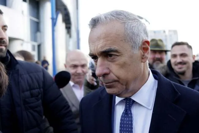 Un bărbat cu părul cărunt, purtând sacou închis și cămașă albastră cu cravată, este înconjurat de alte persoane în exterior.