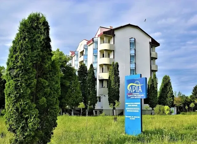 Un clădire albă cu balcone este situată în spatele unor copaci înalți și verzi, lângă un panou informativ albastru.