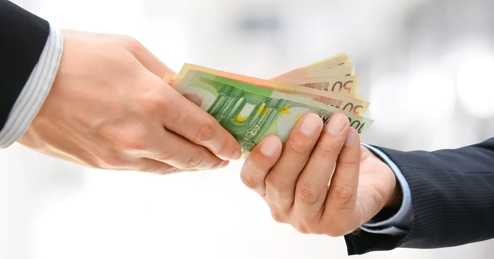 Două mâini, îmbrăcate în manșete de sacou, transmit un teanc de bancnote de euro pe un fundal alb și încețoșat.