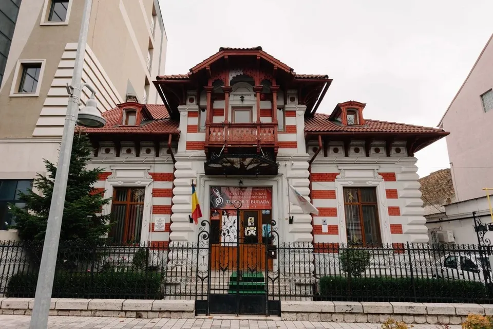 O clădire cu fațadă albă și roșie, având un acoperiș înclinat și ferestre cu rame maronii, este înconjurată de un gard metalic negru.