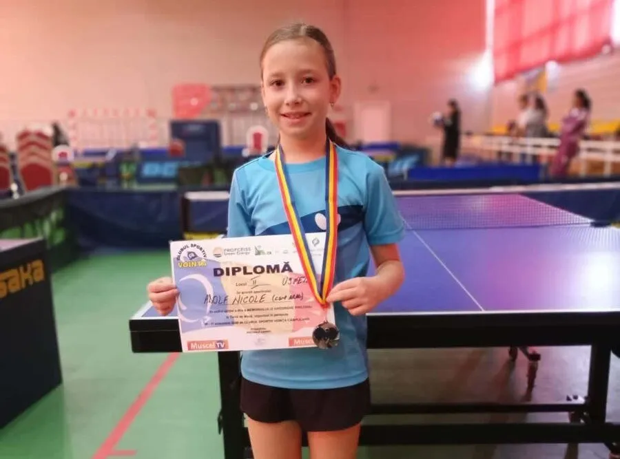 O fată cu tricou albastru și medalie la gât ține o diplomă în fața unei mese de tenis de masă într-o sală de sport.