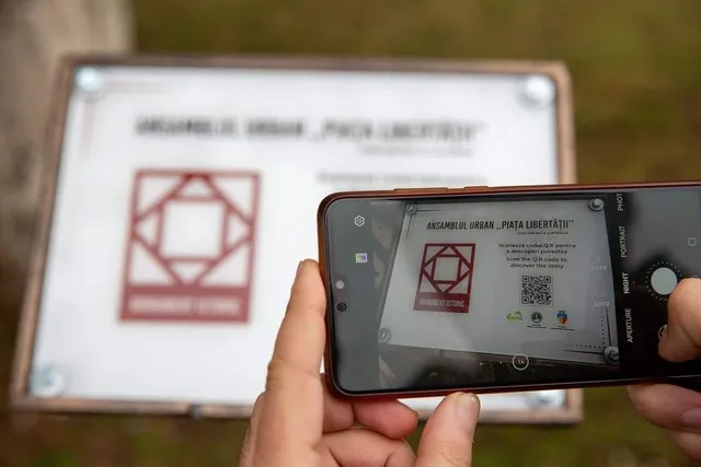 O persoană ține un telefon mobil în poziție orizontală, capturând o placă informativă albă cu un simbol roșu și un cod QR.