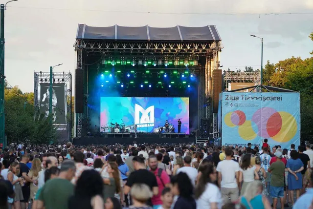 O mulțime de oameni stă în picioare în fața unei scene de concert cu lumini colorate și un ecran mare afișând un logo alb.