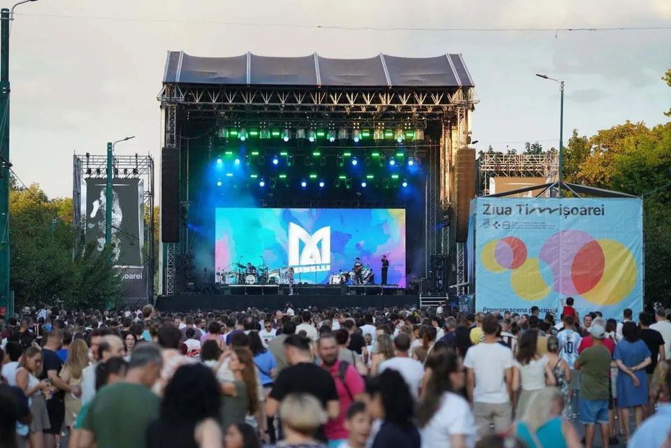 O mulțime de oameni stă în picioare în fața unei scene de concert cu lumini colorate și un ecran mare afișând un logo alb.