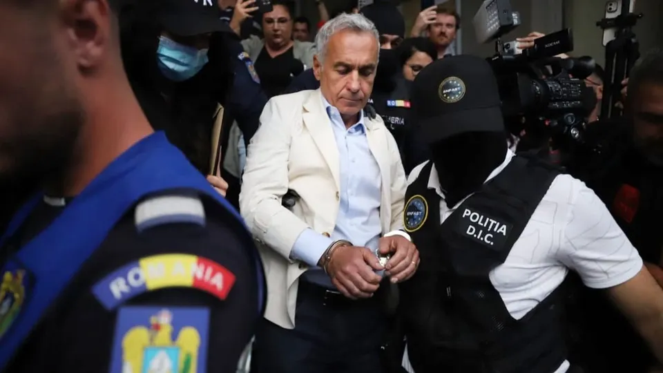 Un bărbat cu părul cărunt și sacou deschis merge printre polițiști și persoane care filmează într-un spațiu aglomerat.