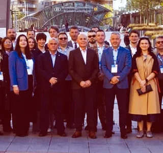 Un grup de persoane îmbrăcate în haine office stă în picioare în fața unei structuri metalice de sticlă sub un cer luminos.