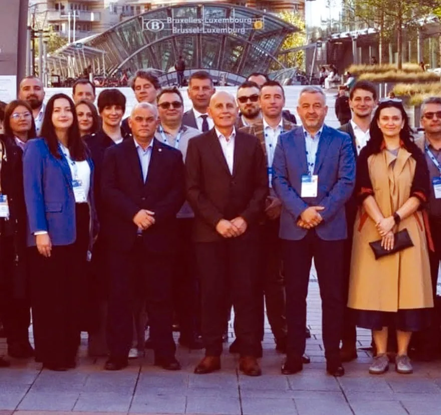 Un grup de persoane îmbrăcate în haine office stă în picioare în fața unei structuri metalice de sticlă sub un cer luminos.