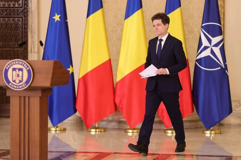 Un bărbat într-un costum închis și cravată merge pe un podea de marmură purtând hârtii albe, în fața mai multor steaguri colorate.