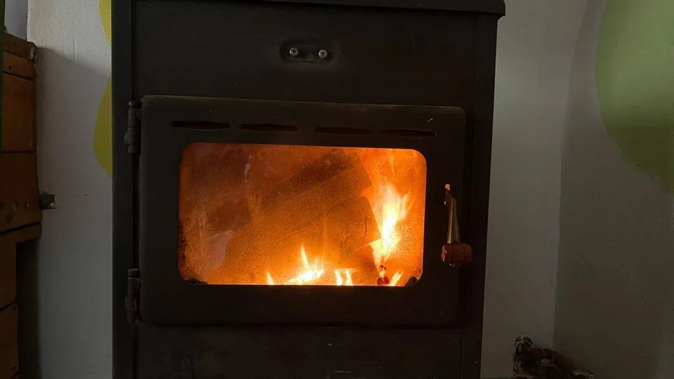 Un foc arde în interiorul unei cuptoare sau șemineu metalice închise într-un cadru interior.