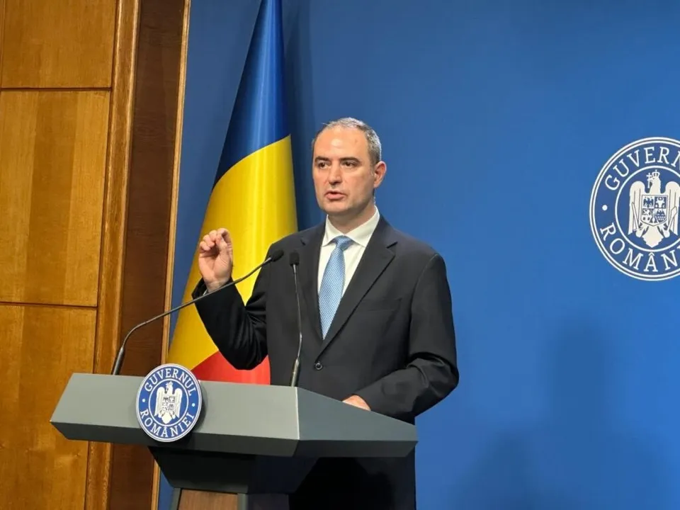 Un bărbat într-un costum închis și cravată deschisă stă în spatele unui podium gri în fața unui steag și a unui fundal albastru.