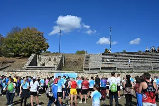 Un grup de oameni stă pe un teren cu iarbă și trepte de piatră sub un cer albastru cu câteva nori albi.