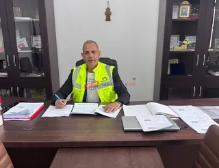 Un bărbat cu vestă reflectorizantă galbenă stă la un birou de lemn în fața unor rafturi cu obiecte și dosare deschise.