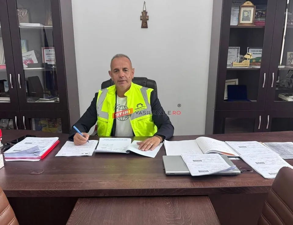 Un bărbat cu vestă reflectorizantă galbenă stă la un birou de lemn în fața unor rafturi cu obiecte și dosare deschise.
